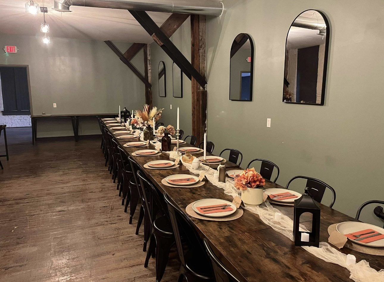 A long dark wood banquet table set with plates, candles, and flowers, with arched mirrors on the sage wall.
