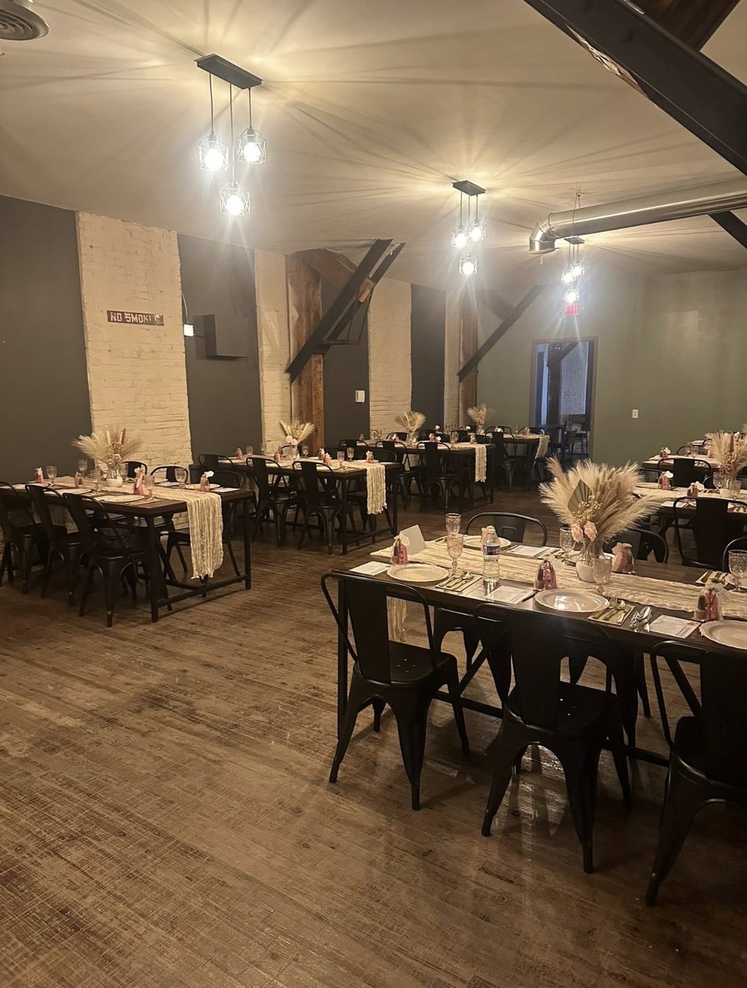 Dining tables dressed with runners and dried grasses in the main room, black chairs, timber posts, and brick.