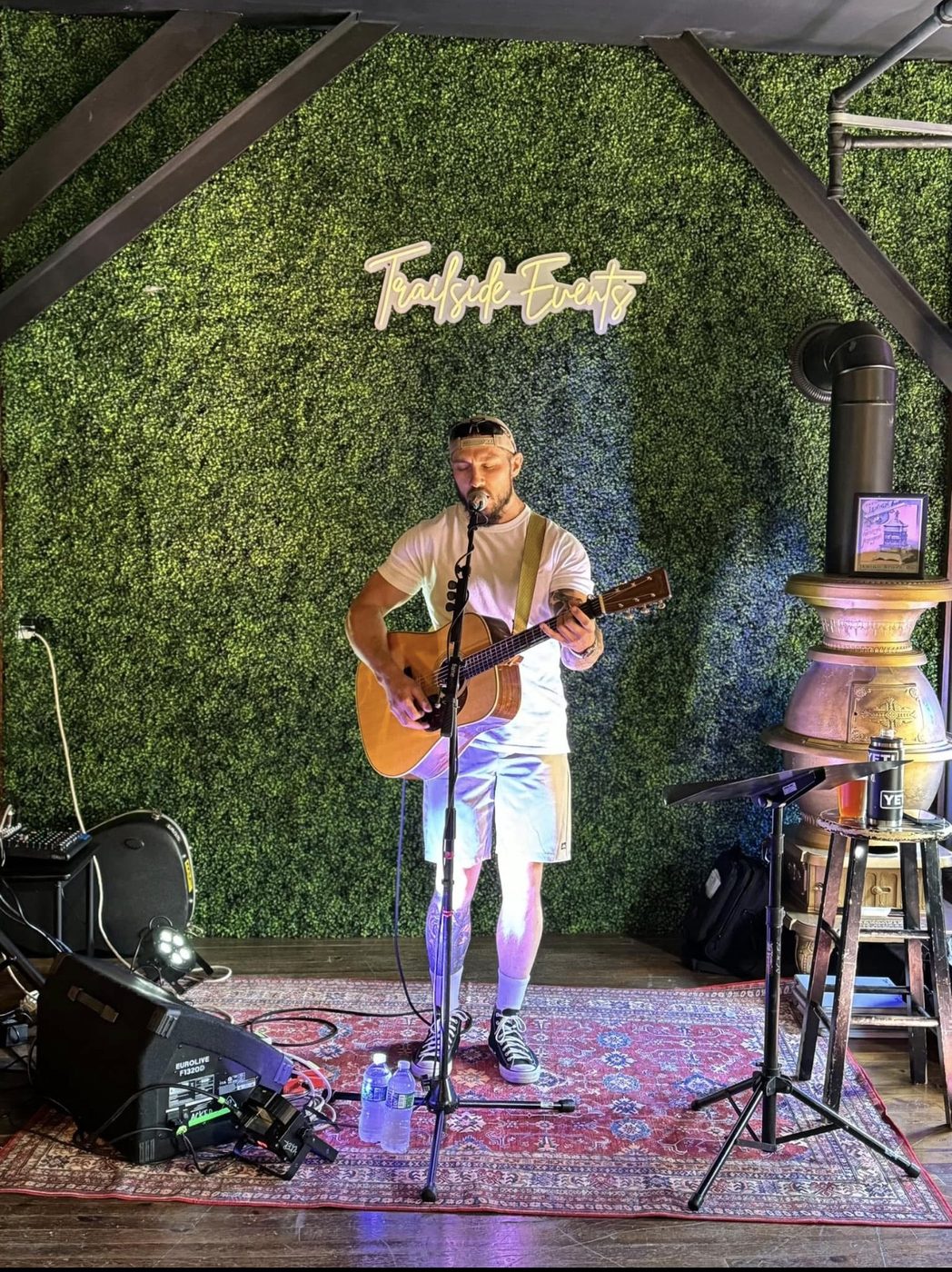 A musician with an acoustic guitar in front of the green wall and TrailSide Events neon, beside the Lehigh stove.